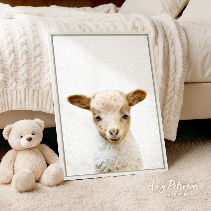 A framed image of a lamb peeking out from behind a white blanket, accompanied by a teddy bear on a beige carpet.