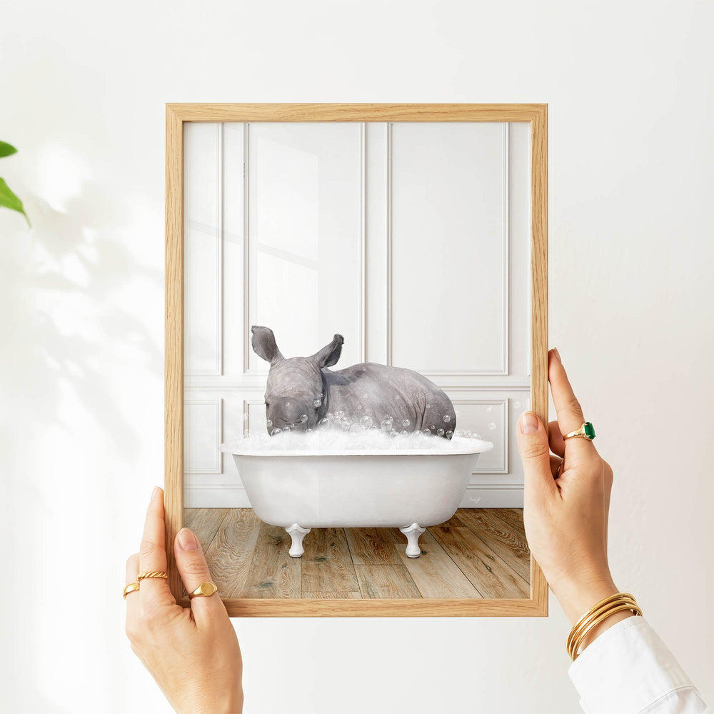 A person's hands are holding a picture frame with a framed image of a rhinoceros sitting in a bathtub filled with water.
