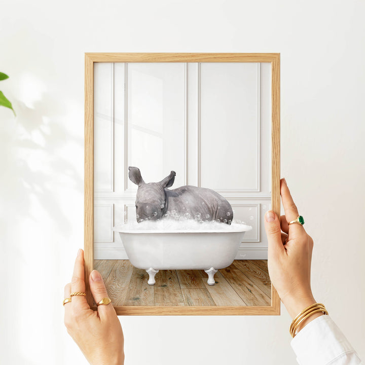 A person's hands are holding a picture frame with a framed image of a rhinoceros sitting in a bathtub filled with water.