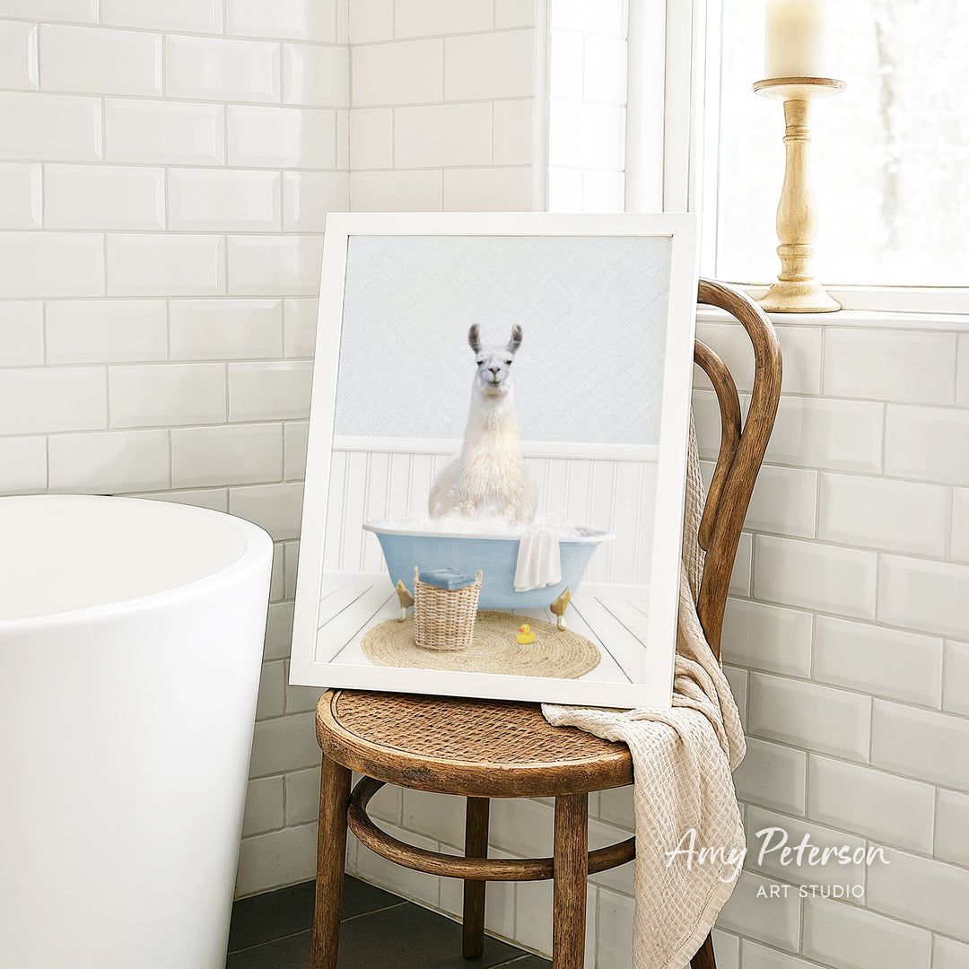 A framed image of a white llama sitting in a blue bathtub, surrounded by a basket of bath toys, is displayed on a wooden chair in a bathroom setting.