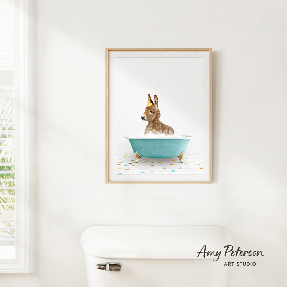 A framed print of a donkey sitting in a blue bathtub, with a colorful tile floor beneath it, is hanging on a white wall.