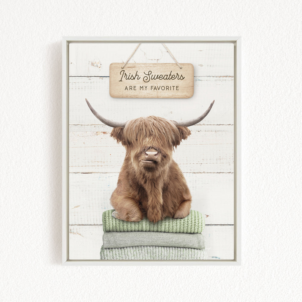 A brown cow with long horns is sitting on a stack of green blankets, with a wooden sign above it that says "Irish Sweaters are my favorite".