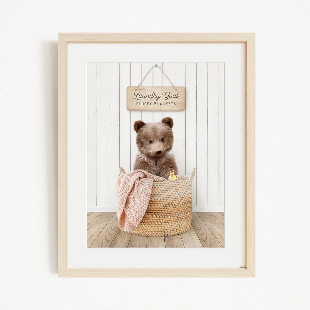 A framed image of a brown bear sitting in a woven basket, with a sign above the basket that reads "Laundry Goal Fluffy Blankets".