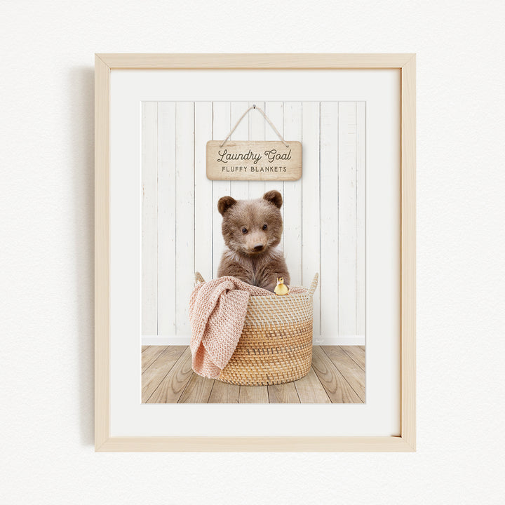 A framed image of a brown bear sitting in a woven basket, with a sign above the basket that reads "Laundry Goal Fluffy Blankets".