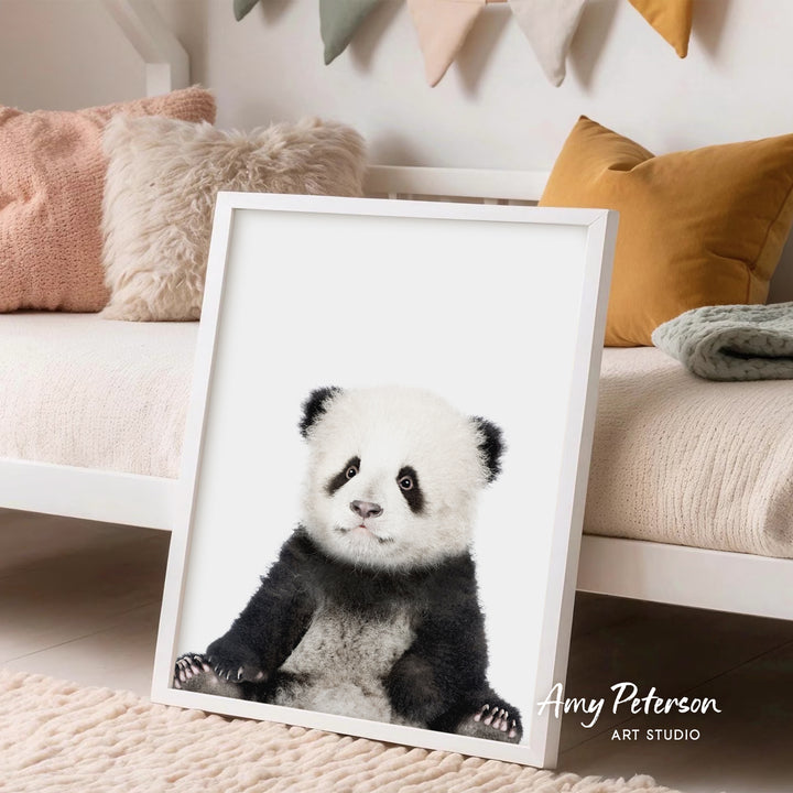 A framed print of a panda bear is displayed in a room with a white bed and colorful pillows in the background.