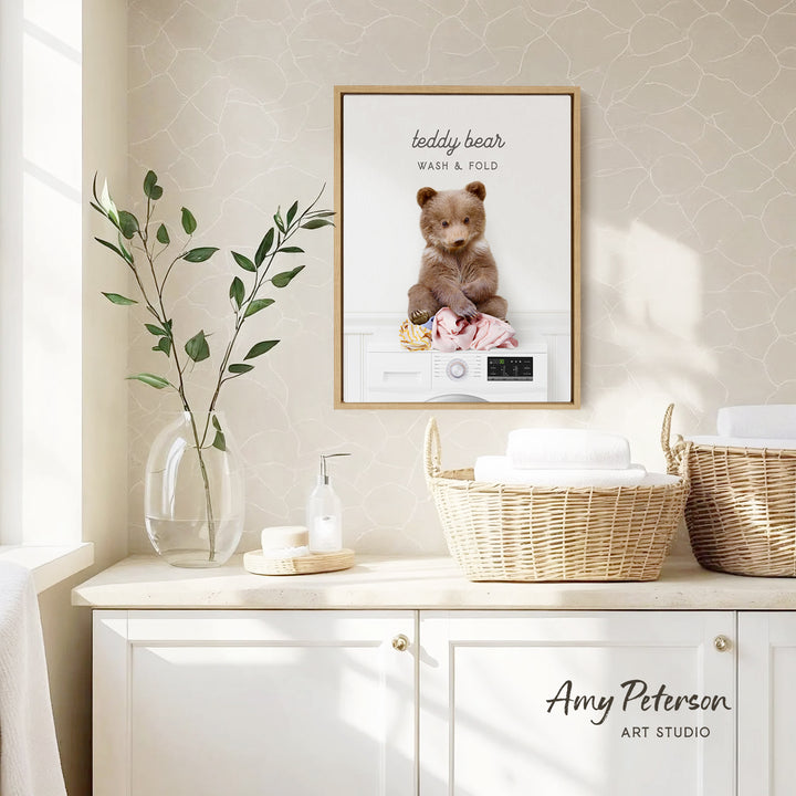 a framed picture of a teddy bear sitting on a washing machine, surrounded by laundry, placed on a wall above a white cabinet.