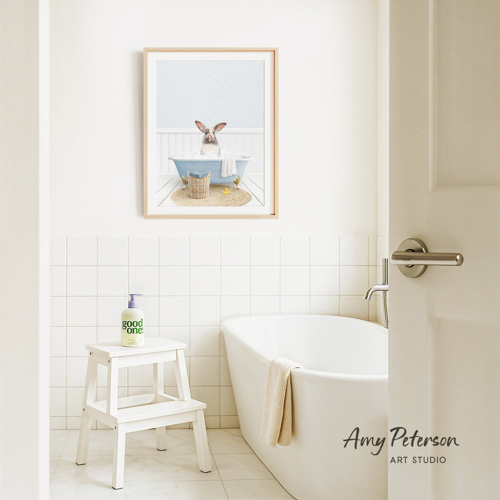A framed image of a rabbit sitting in a bathtub is displayed on the wall above a white bathtub in a bathroom.
