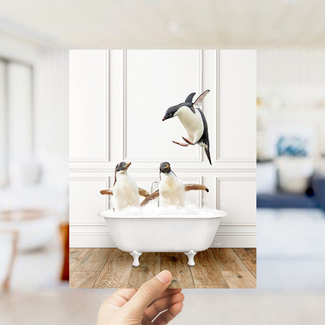 A hand holds up a framed photograph of two penguins sitting in a bathtub, with one penguin jumping out of the tub.