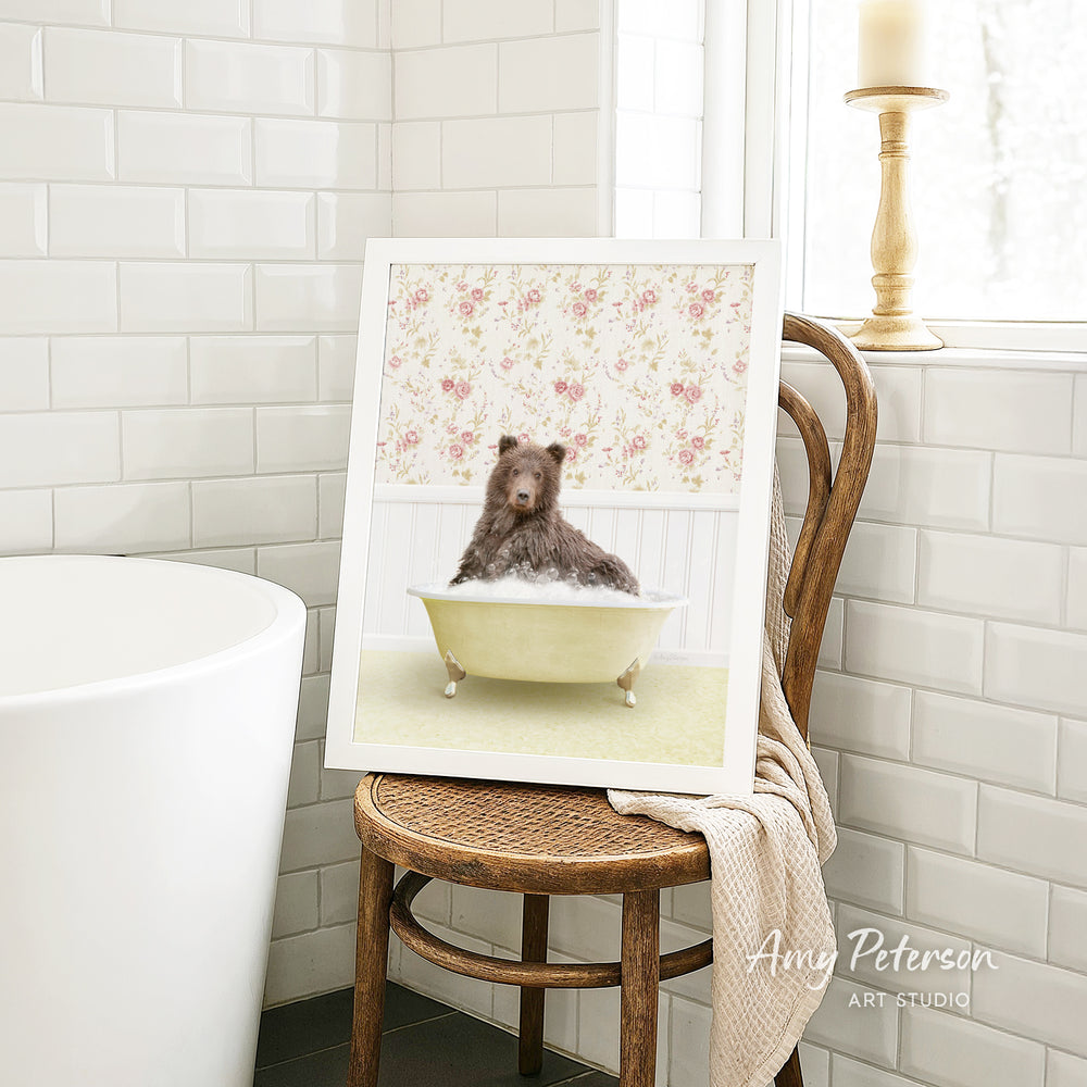 A framed image of a bear sitting in a bathtub, with a floral pattern on the wall behind it, is displayed on a wooden chair in a bathroom setting.