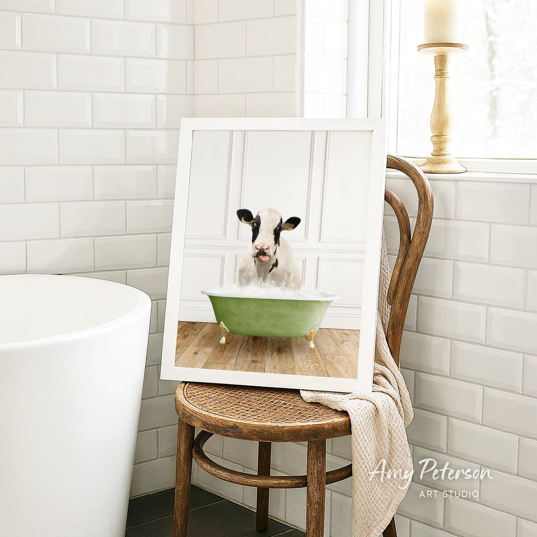 A framed image of a cow sitting in a green bathtub, with a towel draped over the back of a wooden chair in a bathroom setting.