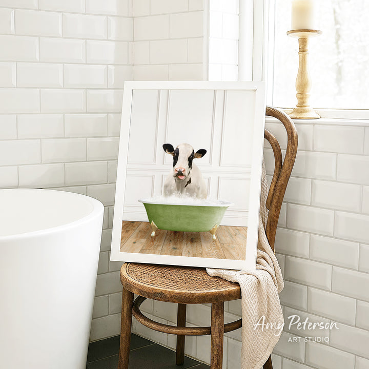 A framed image of a cow sitting in a green bathtub, with a towel draped over the back of a wooden chair in a bathroom setting.