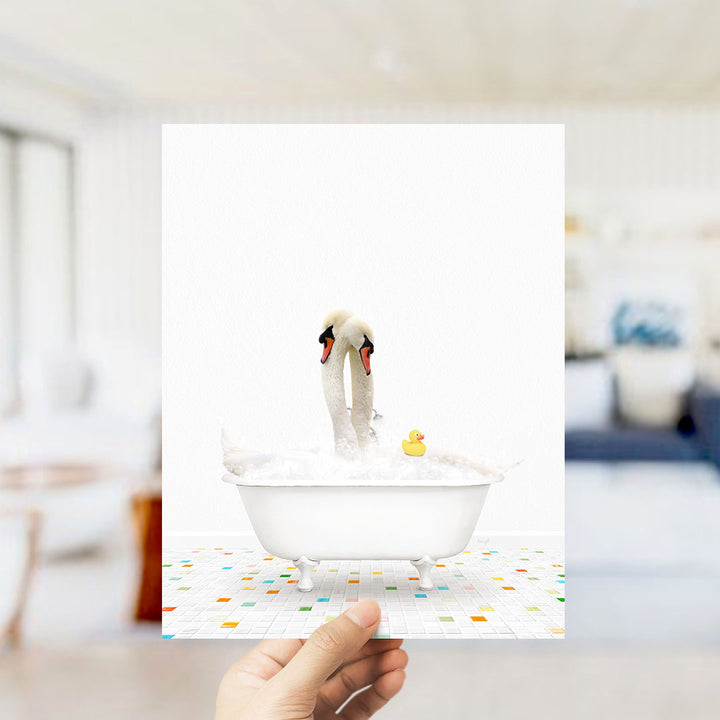 A hand holds up a framed image depicting two swans in a bathtub, with a yellow rubber duck floating in the water.