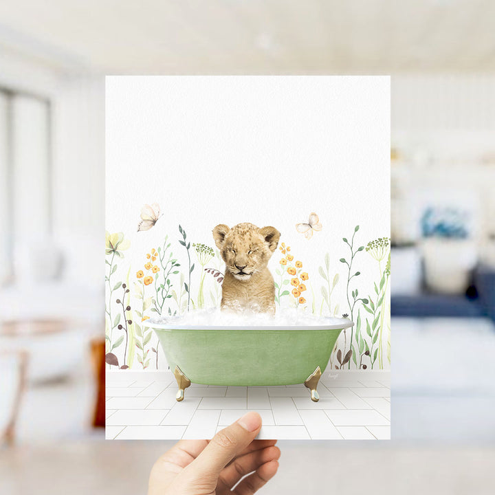 A hand holds up a framed image depicting a young lion cub sitting in a green bathtub filled with bubbles, surrounded by a floral wallpaper.