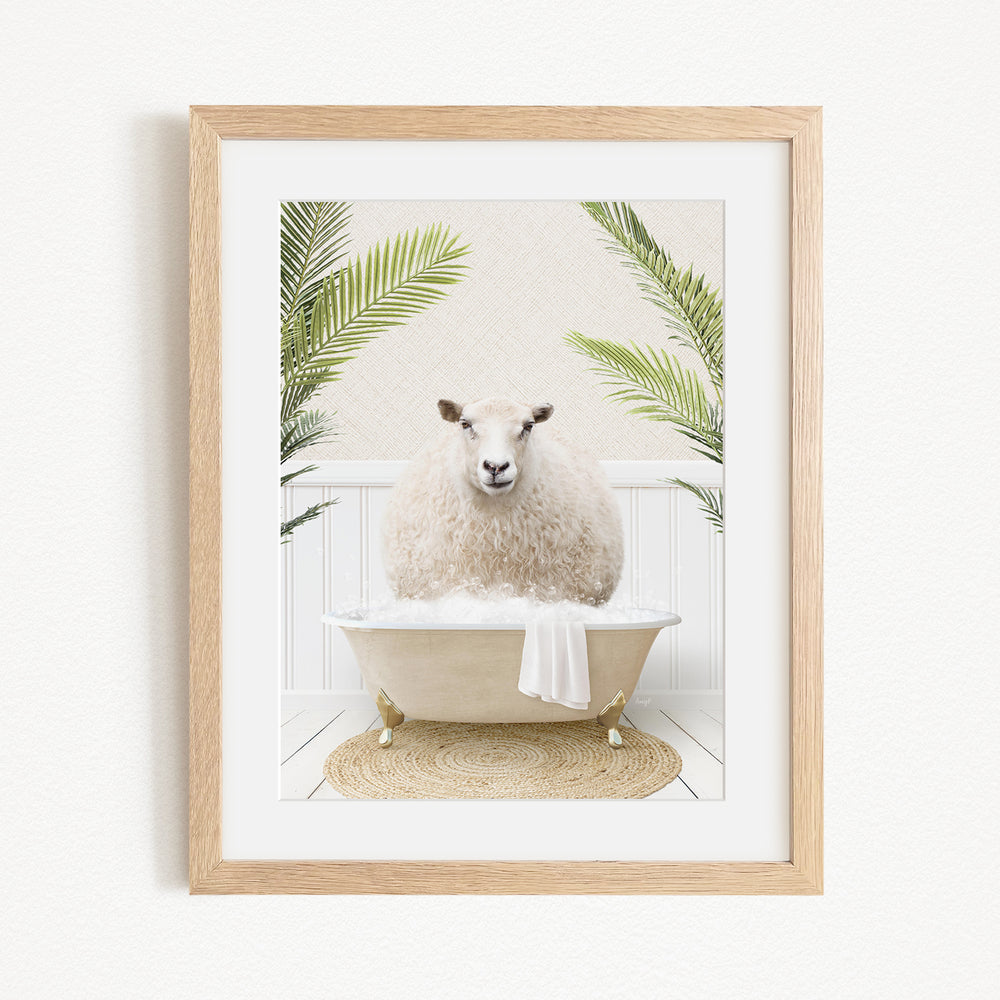 A framed image depicting a fluffy white sheep sitting in a bathtub filled with bubbles, surrounded by green palm fronds.