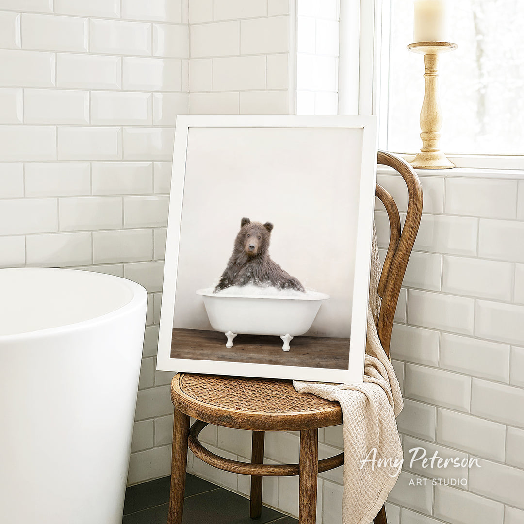 A framed photograph of a bear sitting in a white bathtub is displayed on a wooden chair in a bathroom setting.