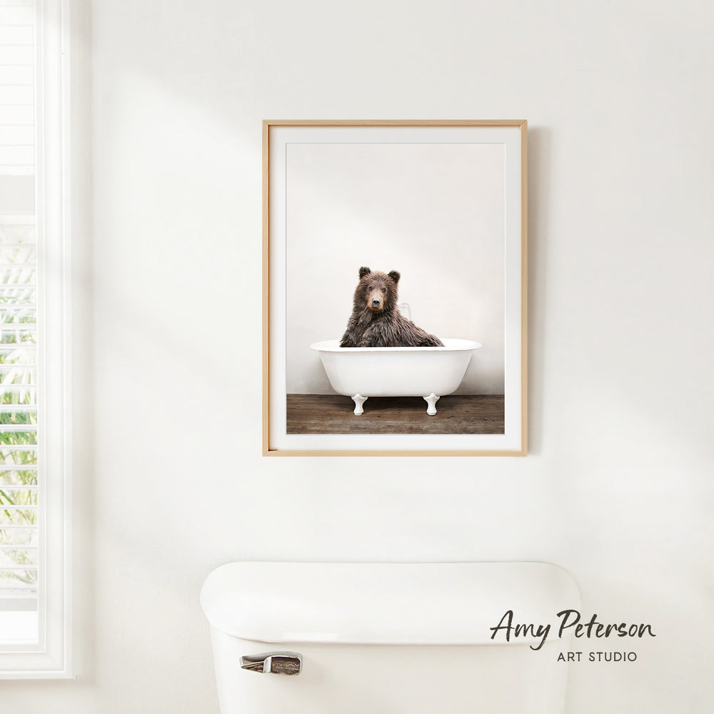 A framed image of a bear sitting in a bathtub is hanging on a white wall.