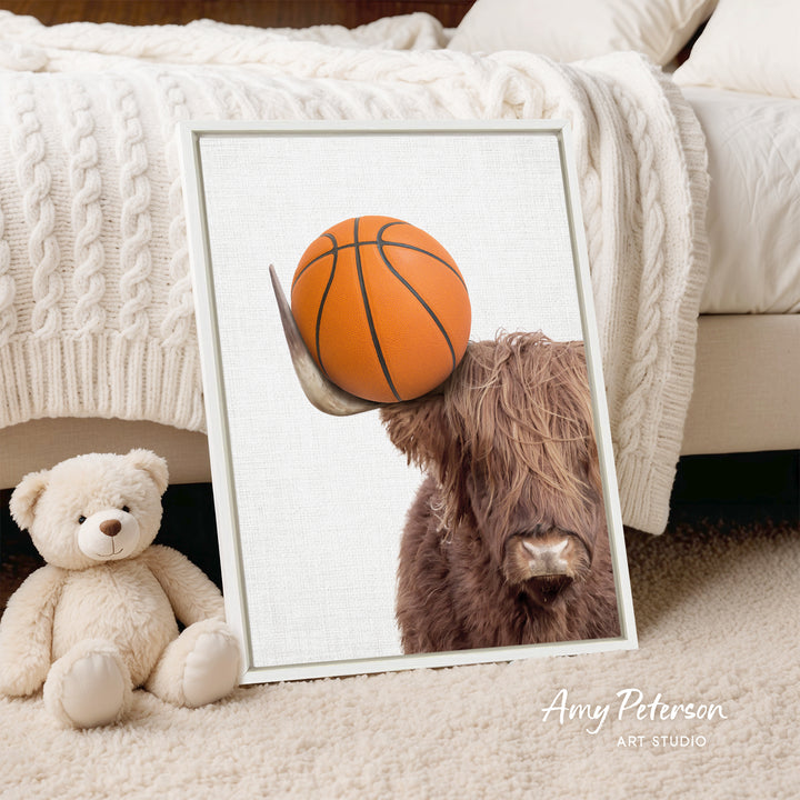 A framed image of a brown cow holding a basketball is displayed on a white bedspread, with a teddy bear next to it.