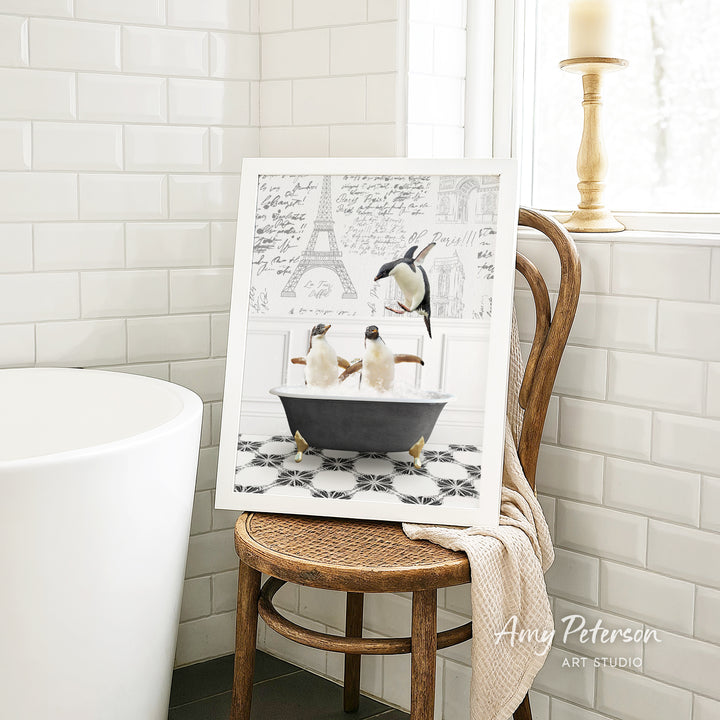 A framed image of two penguins sitting in a bathtub, surrounded by a tiled pattern, is displayed on a wooden chair in a bathroom setting.