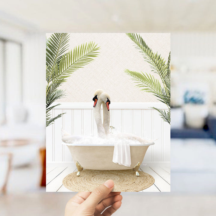 A hand holds up a framed image depicting a pair of swans sitting in a bathtub surrounded by lush green plants.
