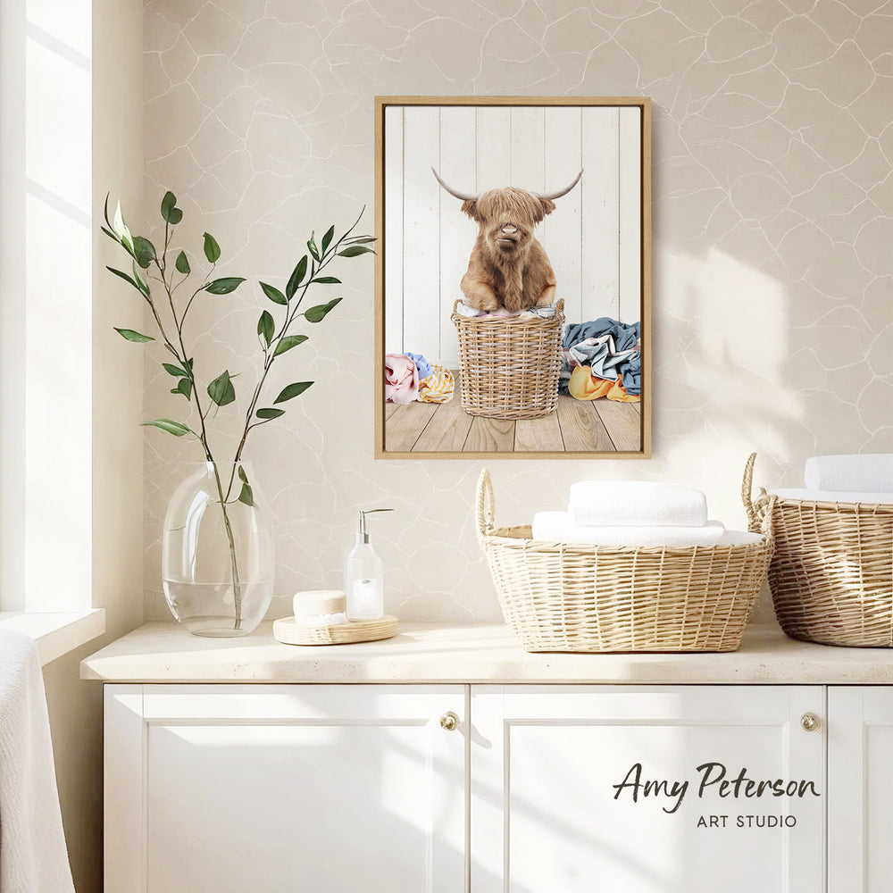 a framed picture of a Highland cow sitting in a wicker basket, placed on a white wall above a white cabinet.