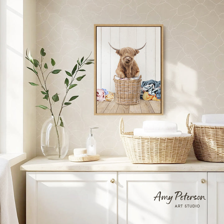 a framed picture of a Highland cow sitting in a wicker basket, placed on a white wall above a white cabinet.