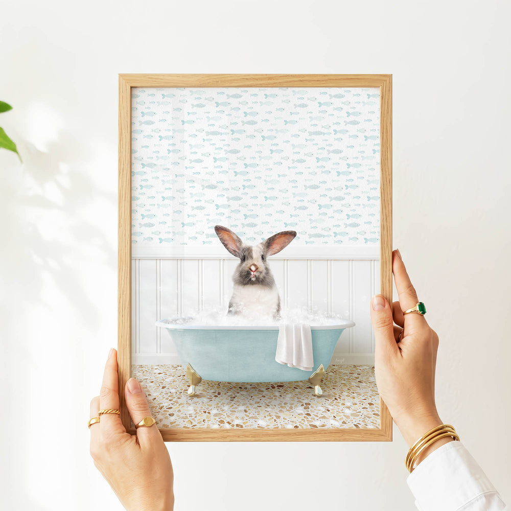 A person's hands are holding a framed picture of a rabbit sitting in a bathtub filled with water, surrounded by a pattern of fish.