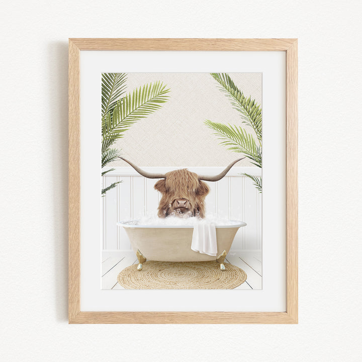 A framed image depicting a Highland cow with long horns sitting in a bathtub filled with bubbles, surrounded by green palm fronds.