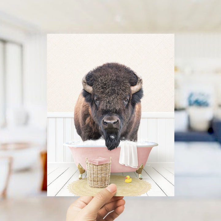 A buffalo with large horns is sitting in a pink bathtub filled with bubbles, surrounded by a yellow rubber duck and a white towel.