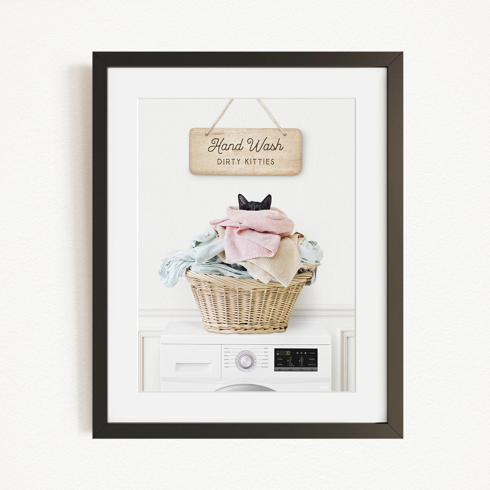 A framed image of a cat sitting in a basket filled with laundry, with a sign above that says "Hand Wash Dirty Kittens".