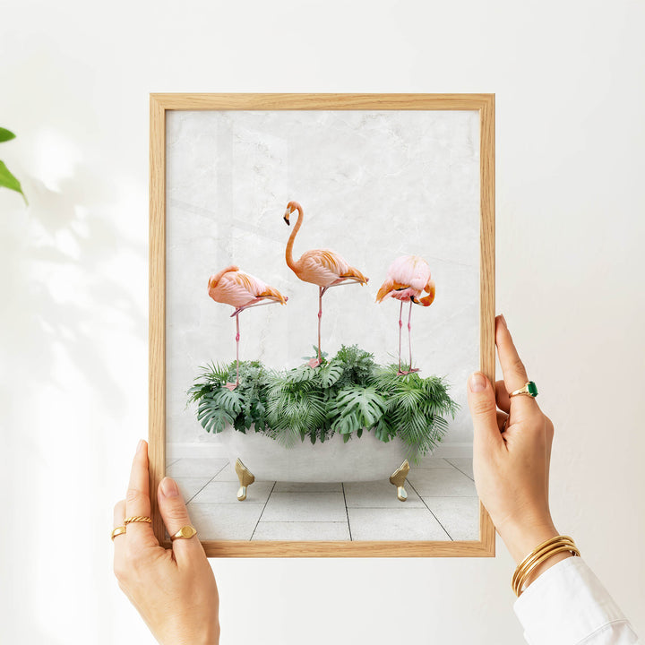 A person's hands are holding a framed picture of three pink flamingos standing on a green plant in a white vase.