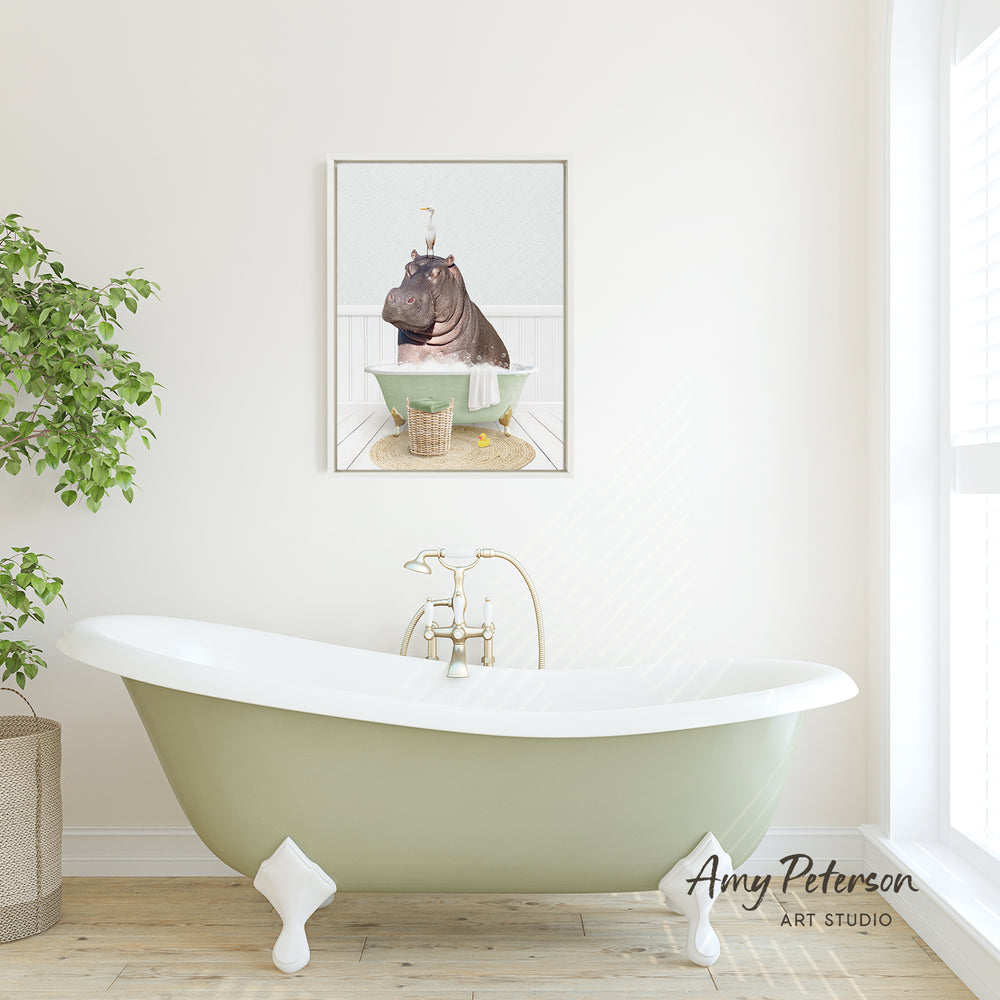 A framed image of a hippopotamus sitting in a bathtub is displayed on the wall above a green clawfoot bathtub in a bright, white-walled bathroom.