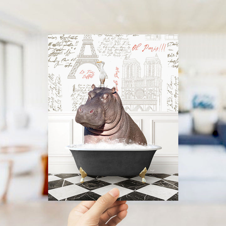 A hand holds up a printed image featuring a hippopotamus sitting in a bathtub, surrounded by various Paris-themed elements such as the Eiffel Tower and the Notre-Dame Cathedral.