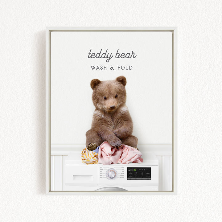 A teddy bear sitting on a white washing machine, with a pile of laundry next to it.