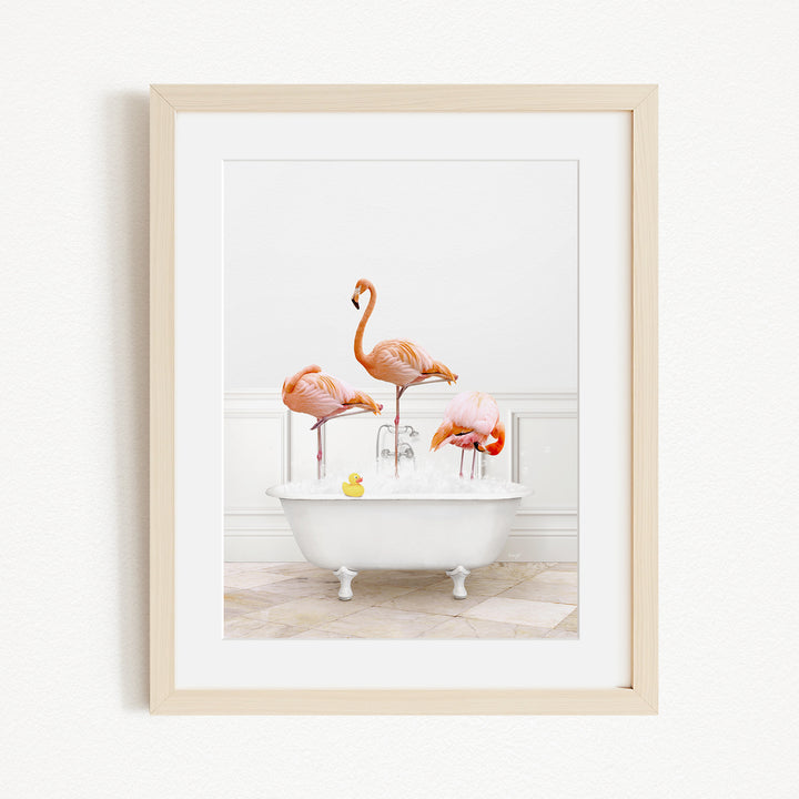 A framed image depicting three flamingos standing in a white bathtub filled with water, with a yellow rubber duck floating in the water.