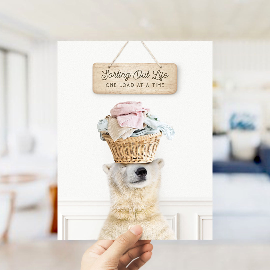 A polar bear wearing a basket of laundry and looking at the camera, with a wooden sign above that says "Sorting Out Life One Load at a Time".