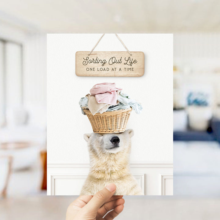 A polar bear wearing a basket of laundry and looking at the camera, with a wooden sign above that says "Sorting Out Life One Load at a Time".