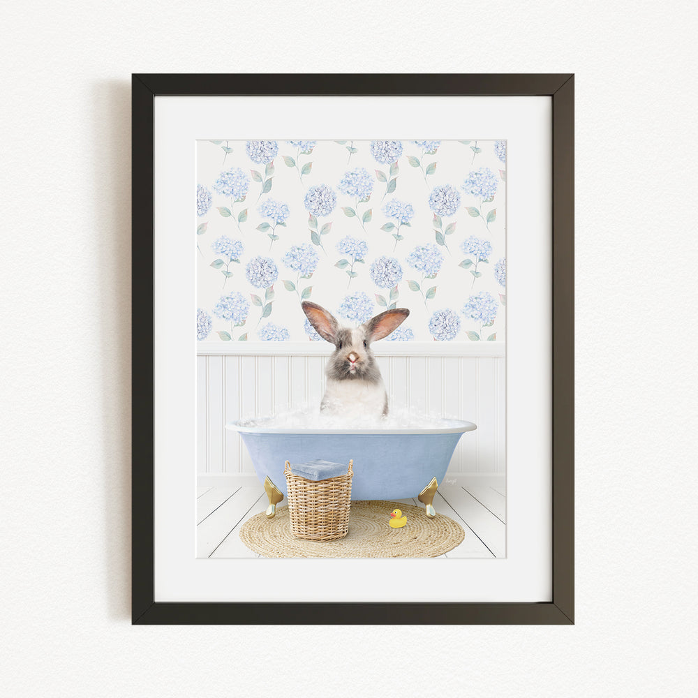 A rabbit sitting in a blue bathtub filled with water, surrounded by a floral patterned wall and a basket of rubber ducks.