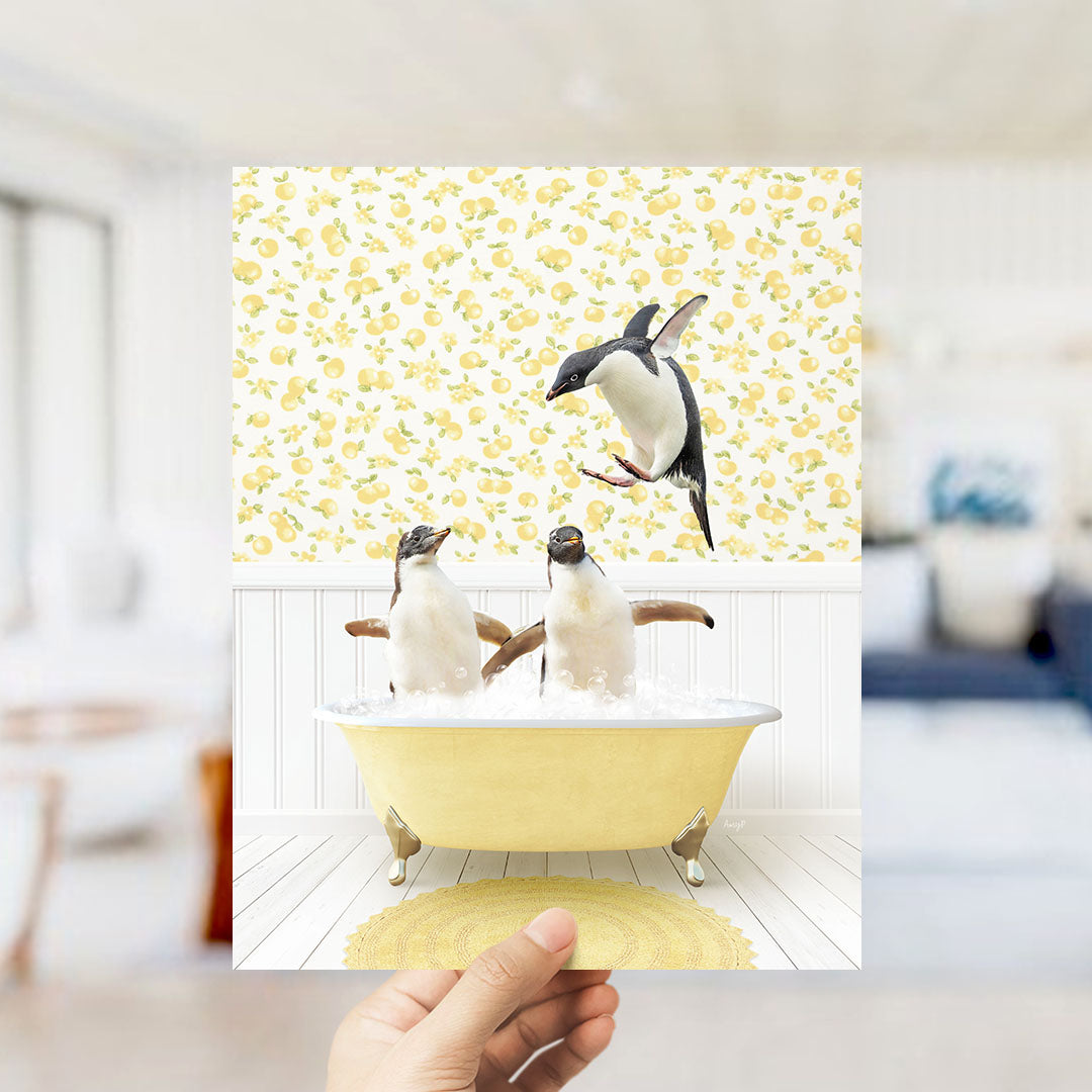 A hand holds up a framed image depicting two penguins sitting in a yellow bathtub, with one penguin jumping out of the tub.