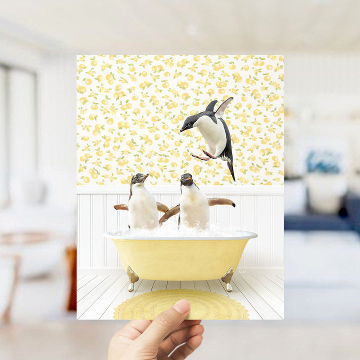 A hand holds up a framed image depicting two penguins sitting in a yellow bathtub, with one penguin jumping out of the tub.