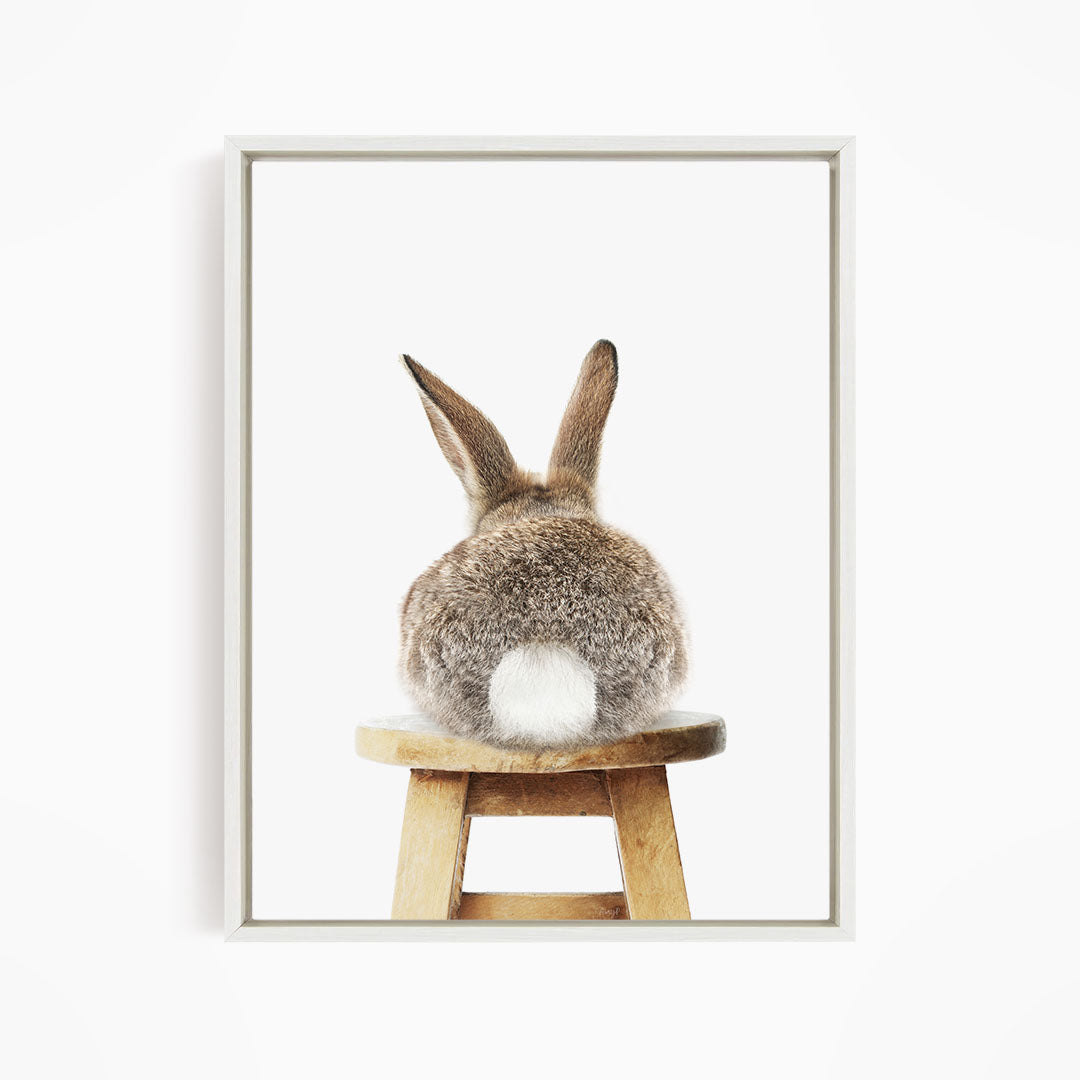 A brown hare sits on a wooden stool, facing away from the camera.