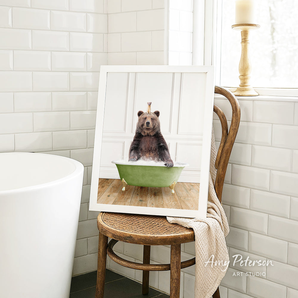 A framed image of a bear sitting in a green bathtub, with a towel draped over the back of the chair.