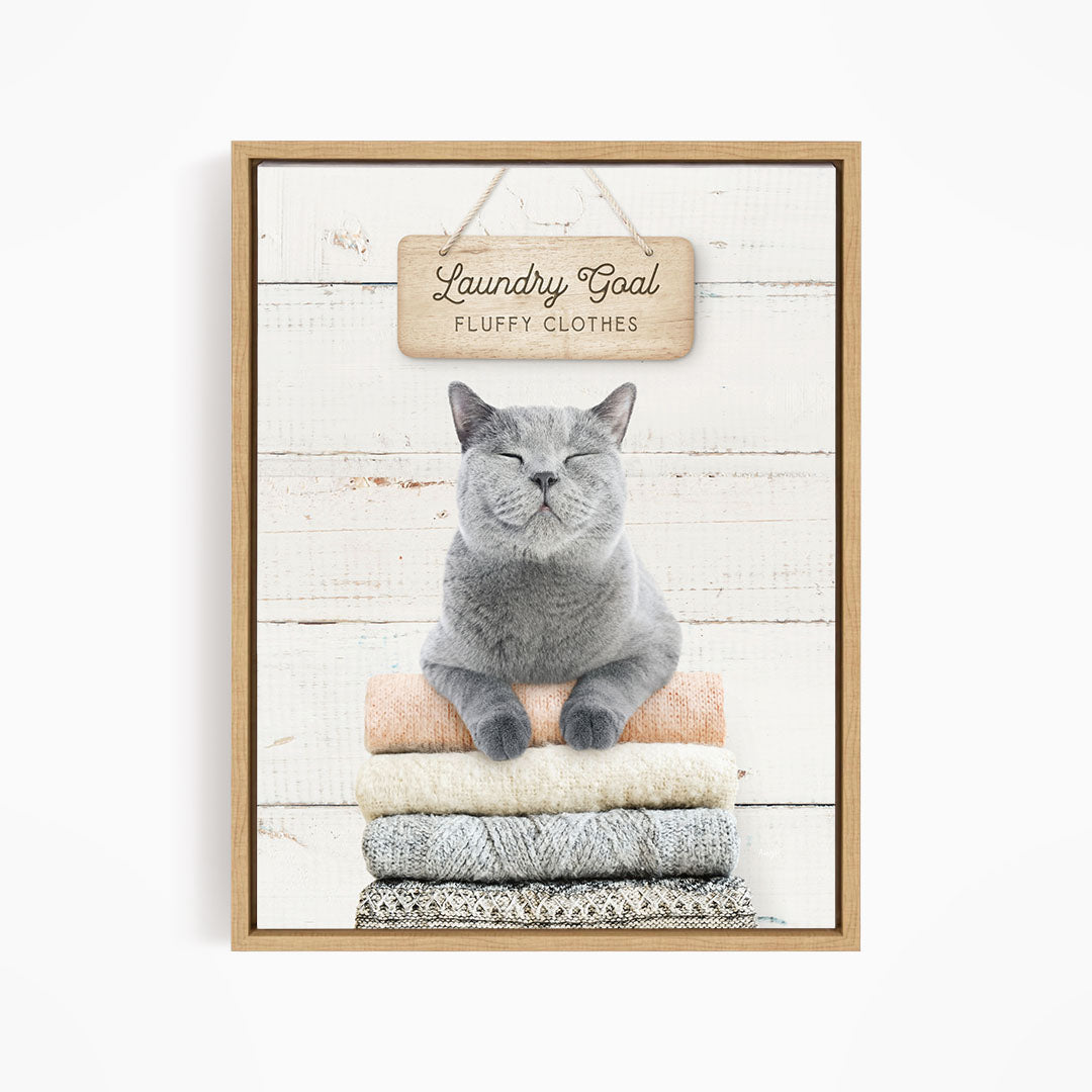 A gray cat sitting on a stack of folded clothes, with a wooden sign above that says "Laundry Goal Fluffy Clothes".