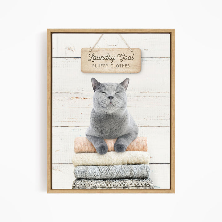 A gray cat sitting on a stack of folded clothes, with a wooden sign above that says "Laundry Goal Fluffy Clothes".