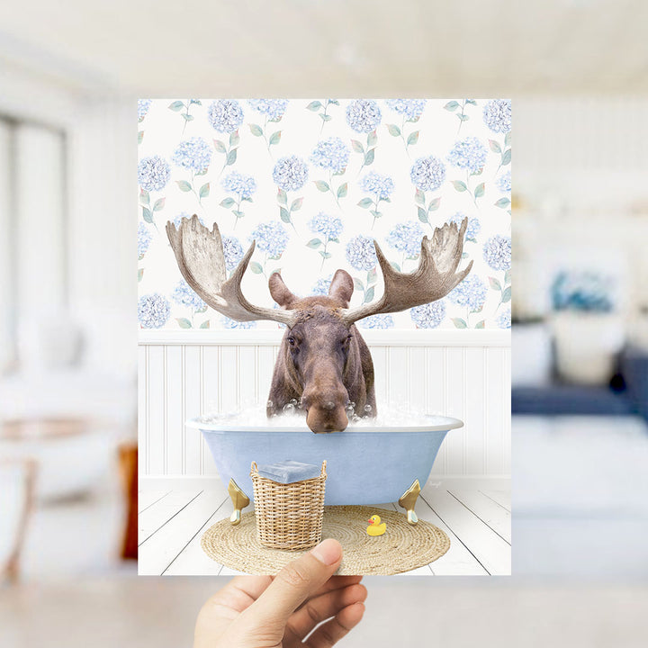 A moose with large antlers is sitting in a blue bathtub filled with water, surrounded by a wicker basket and a rubber duck.