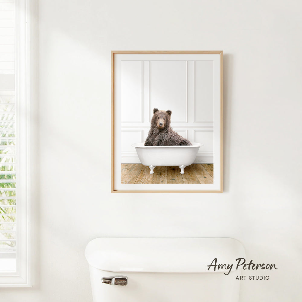 A framed image of a brown bear sitting in a white bathtub is hanging on a white wall.