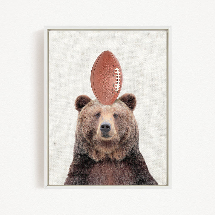 A brown bear with a football on its head, looking directly at the camera.