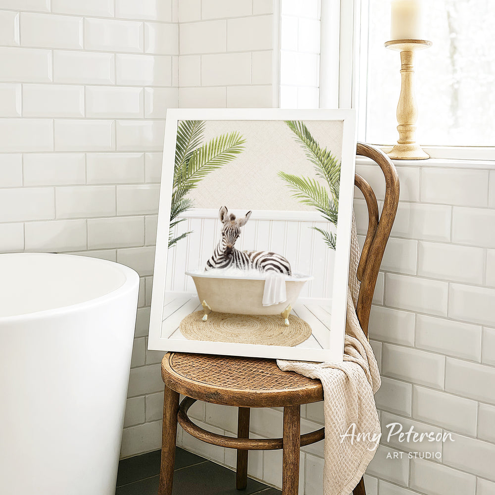 A framed image of a zebra sitting in a bathtub, surrounded by palm leaves, is displayed on a wooden chair in a bathroom setting.