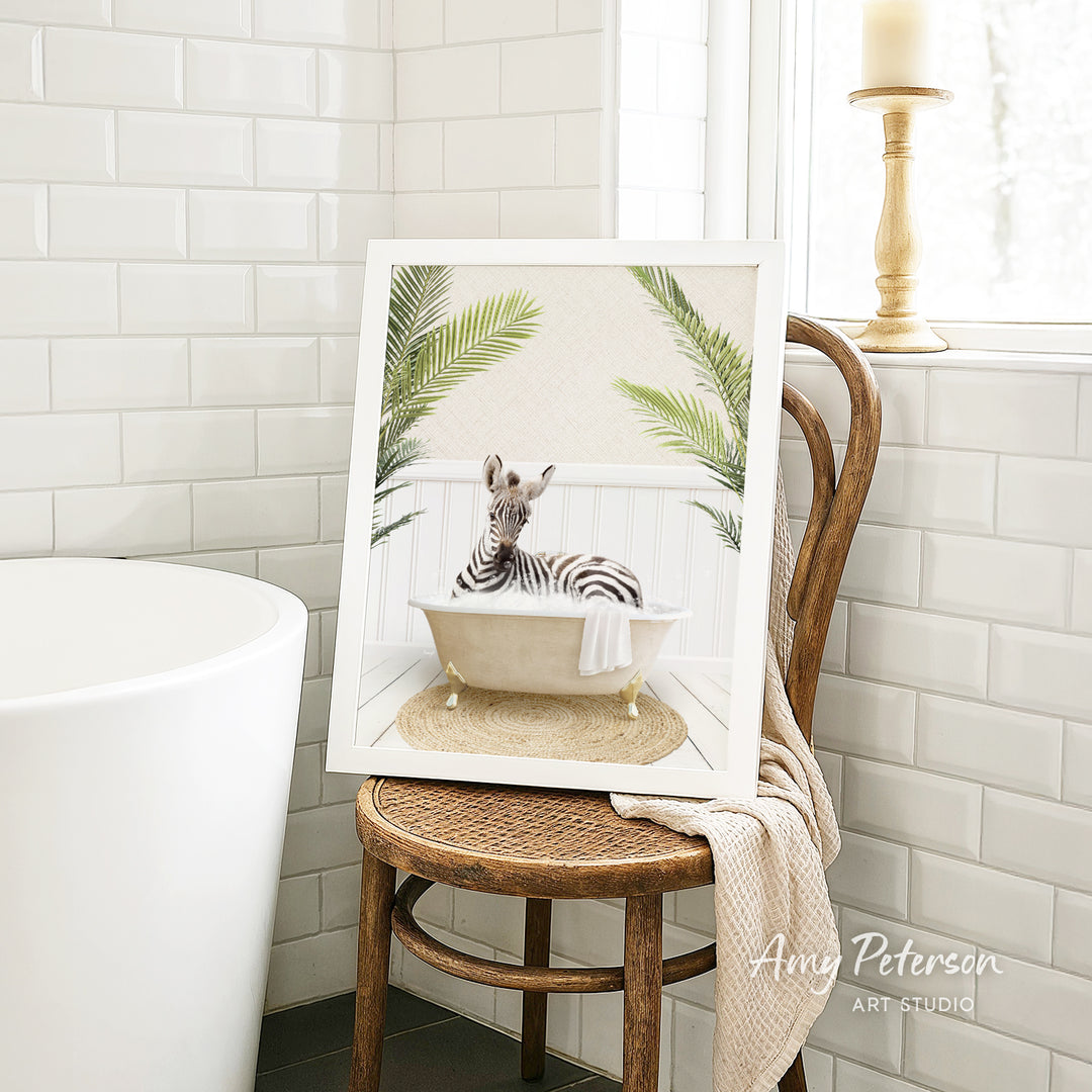 A framed image of a zebra sitting in a bathtub, surrounded by palm leaves, is displayed on a wooden chair in a bathroom setting.