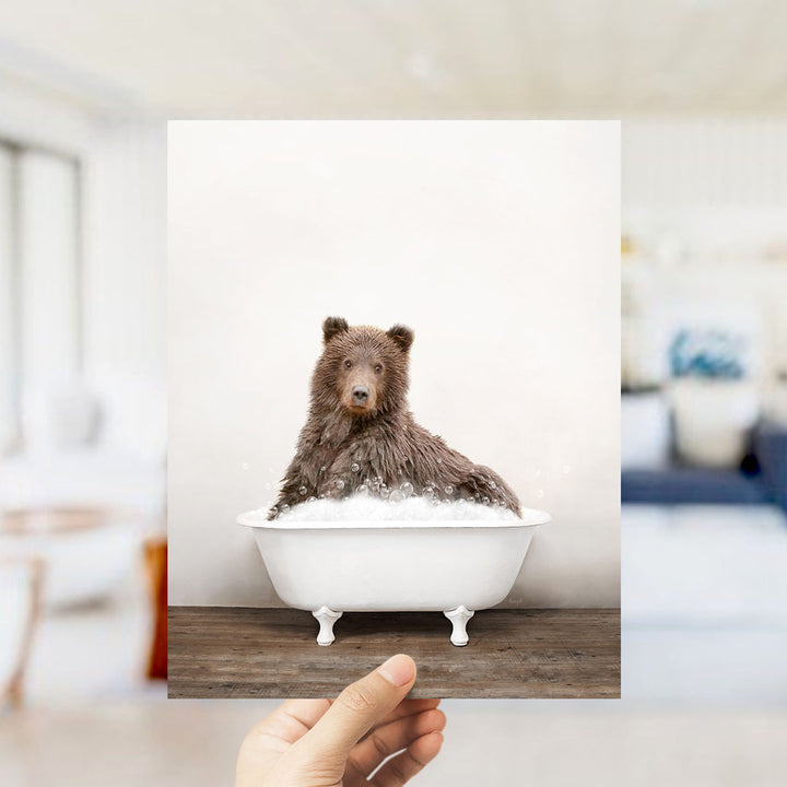 A hand holds up a framed image of a brown bear sitting in a white bathtub filled with bubbles.