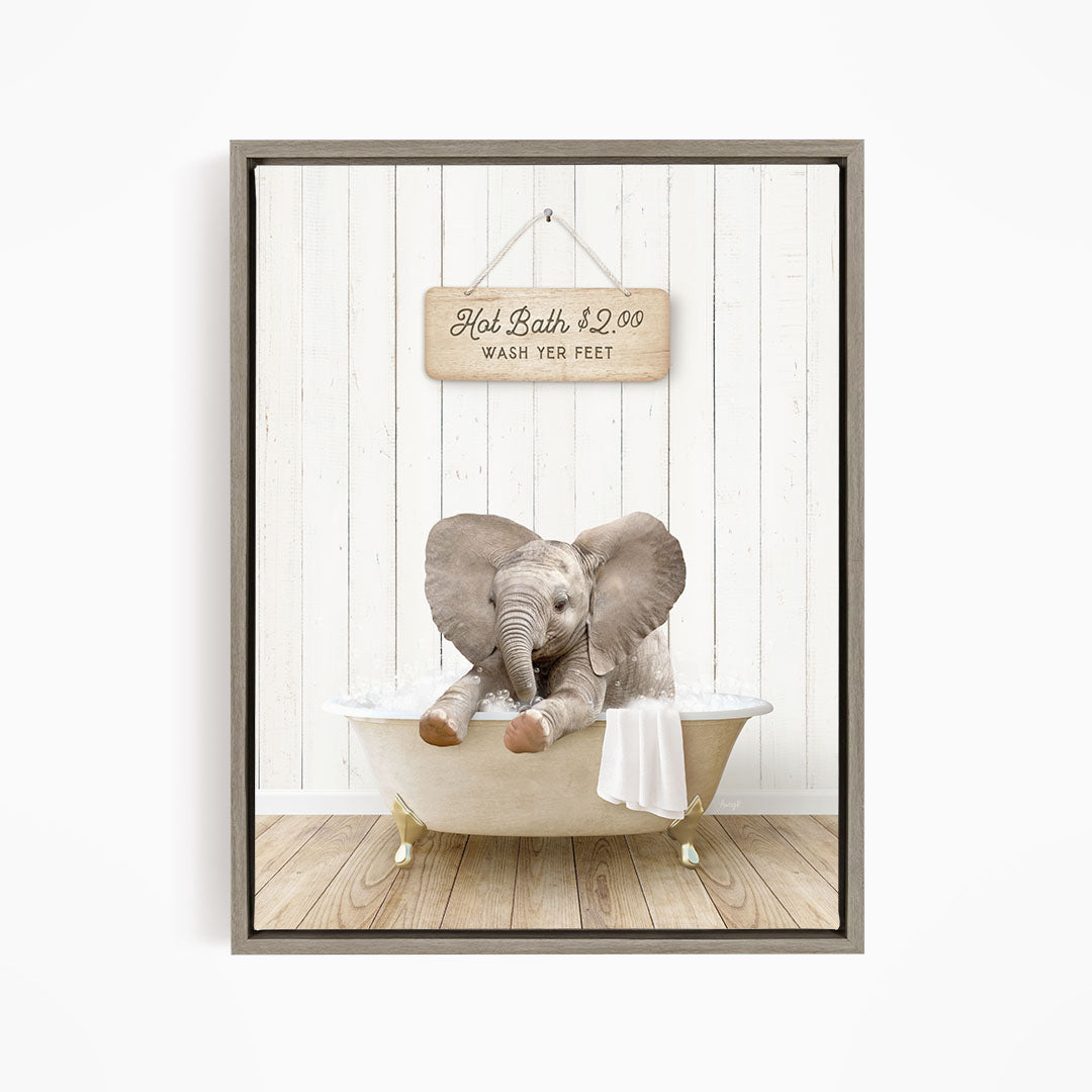 A wooden sign with the text "Hot Bath $2.00 WASH YOUR FEET" hangs on a white wooden wall, with a baby elephant sitting in a bathtub filled with water.